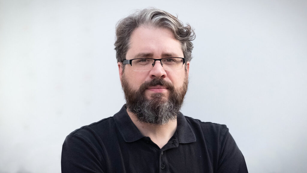 Porträtt på Eliot Higgins i svarta kläder med en grå bakgrund.