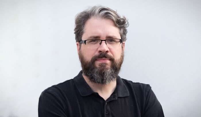 Porträtt på Eliot Higgins i svarta kläder med en grå bakgrund.
