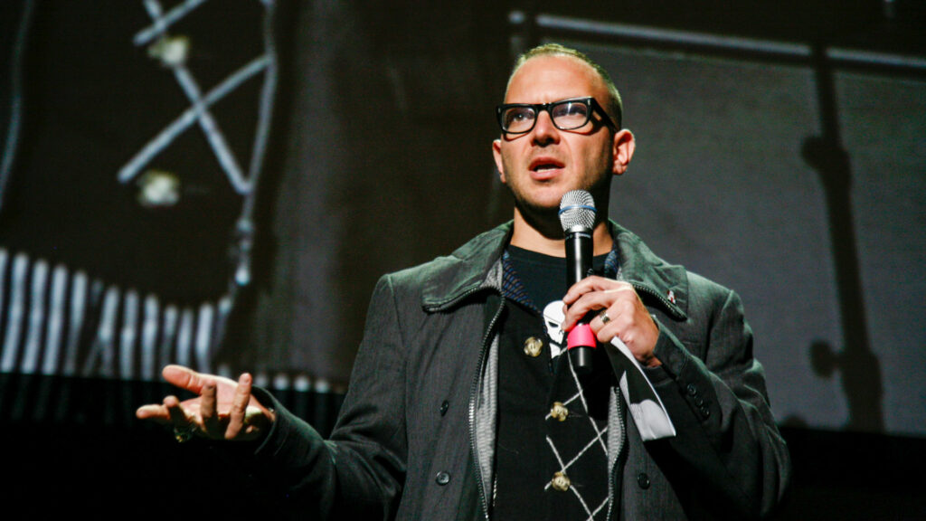 Cory Doctorow talar i en mikrofon på en scen, han gestikulerar med handen.